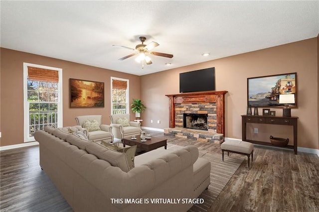 living area featuring dark wood-style floors, a stone fireplace, a ceiling fan, and baseboards