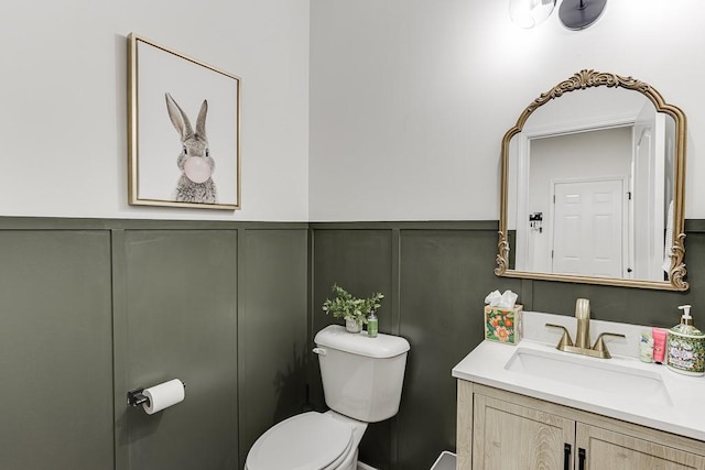 half bathroom featuring a decorative wall, toilet, vanity, and wainscoting