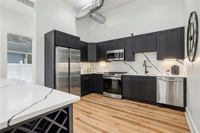 kitchen featuring tasteful backsplash, a towering ceiling, light wood-style flooring, appliances with stainless steel finishes, and a sink