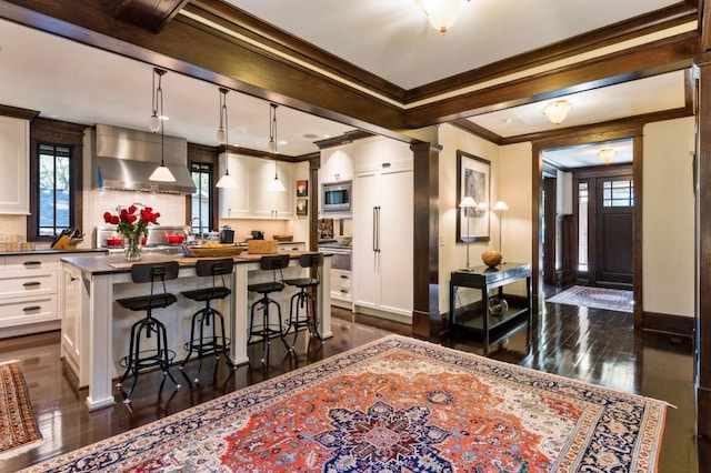 kitchen featuring dark countertops, wall chimney exhaust hood, hanging light fixtures, a kitchen bar, and white cabinetry