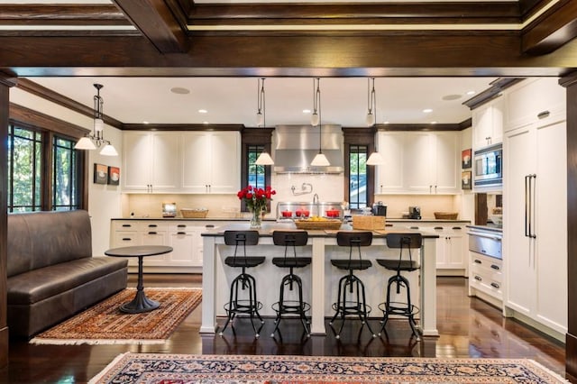 kitchen featuring a kitchen breakfast bar, white cabinets, wall chimney range hood, stainless steel microwave, and decorative light fixtures