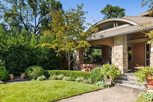 view of exterior entry featuring stone siding, brick siding, a lawn, and roof with shingles