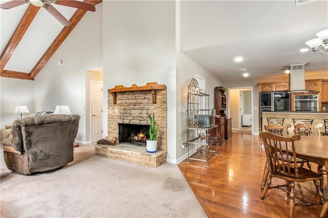 carpeted living room with ceiling fan with notable chandelier, beam ceiling, a stone fireplace, and high vaulted ceiling