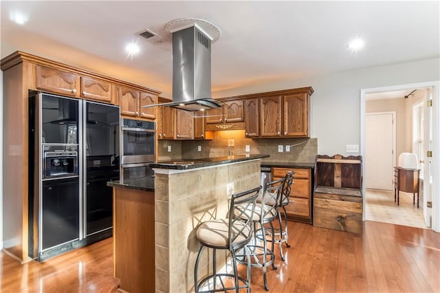 kitchen featuring island exhaust hood, tasteful backsplash, black fridge with ice dispenser, light hardwood / wood-style flooring, and a breakfast bar