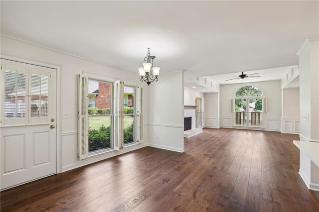 unfurnished living room featuring dark wood finished floors, ornamental molding, a brick fireplace, a decorative wall, and ceiling fan with notable chandelier