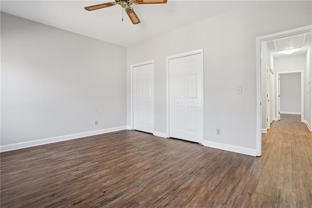 unfurnished bedroom with two closets, baseboards, dark wood finished floors, attic access, and a ceiling fan