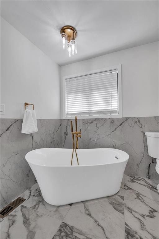 full bathroom featuring tile walls, wainscoting, a soaking tub, marble tiled flooring, and toilet