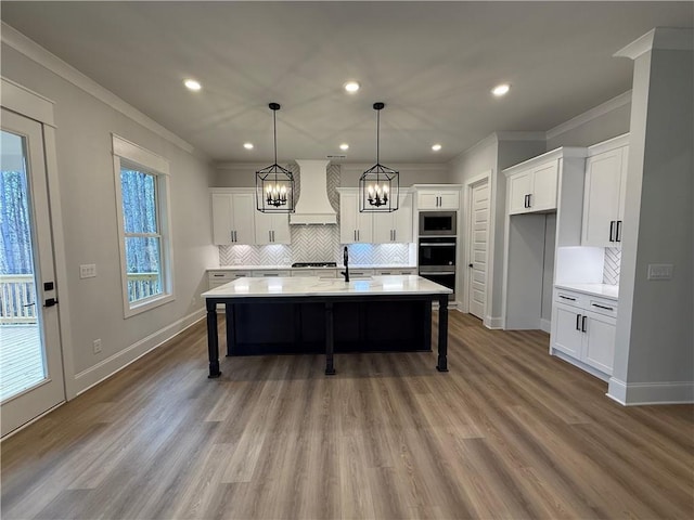 kitchen with crown molding, a kitchen island with sink, custom range hood, and light wood-type flooring