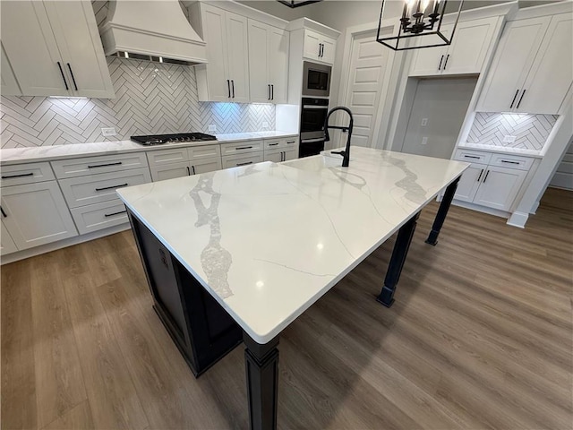 kitchen with backsplash, hanging light fixtures, an island with sink, light stone counters, and custom range hood