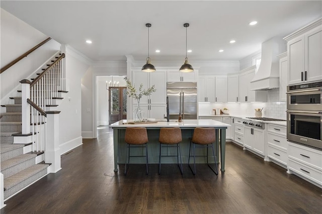 kitchen featuring white cabinetry, stainless steel appliances, custom range hood, a center island with sink, and decorative light fixtures