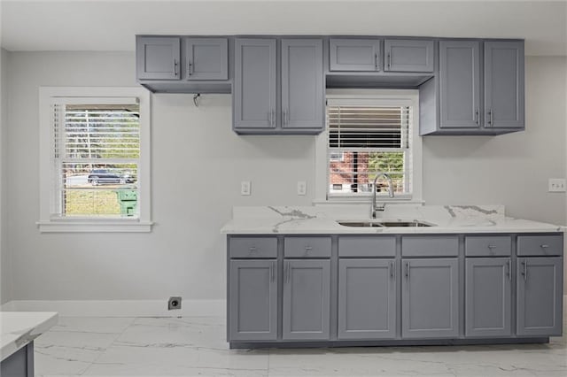 kitchen with baseboards, light stone countertops, gray cabinets, marble finish floor, and a sink