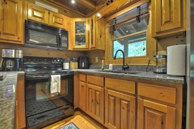 kitchen with a sink, brown cabinetry, dark stone countertops, black appliances, and glass insert cabinets