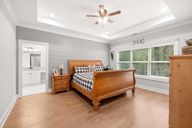 bedroom featuring ensuite bath, wood walls, light hardwood / wood-style floors, a tray ceiling, and ceiling fan