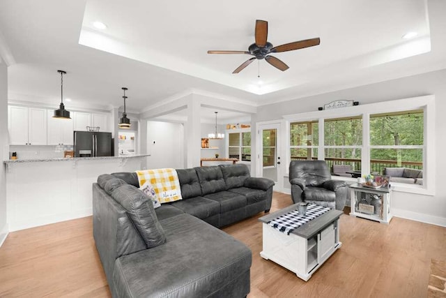 living room with light wood-type flooring, ceiling fan with notable chandelier, a healthy amount of sunlight, and a raised ceiling