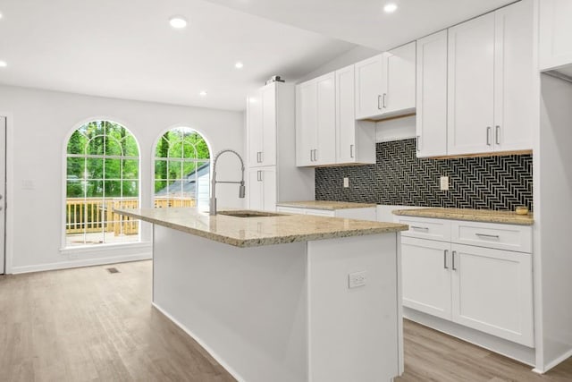 kitchen with tasteful backsplash, light stone countertops, a center island with sink, light wood-type flooring, and a sink