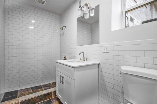 bathroom with vanity, visible vents, tiled shower, tile walls, and toilet