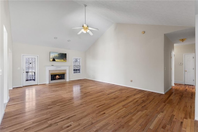 unfurnished living room with ceiling fan, wood finished floors, high vaulted ceiling, a lit fireplace, and baseboards