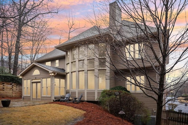 property exterior at dusk with a patio area, fence, a chimney, and a shingled roof