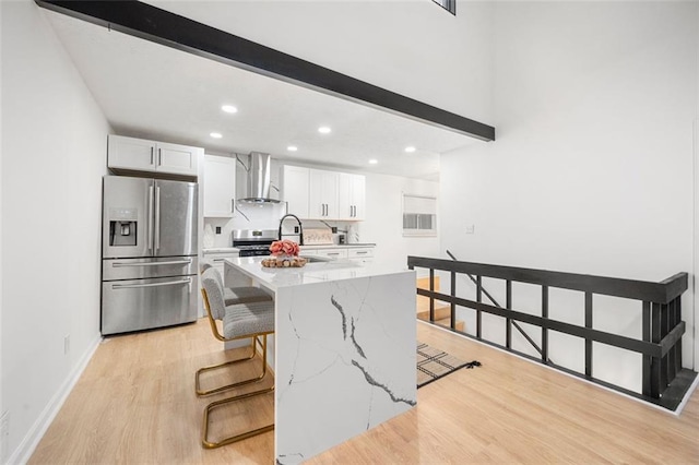 kitchen featuring white cabinetry, a kitchen breakfast bar, stainless steel appliances, light stone countertops, and wall chimney range hood