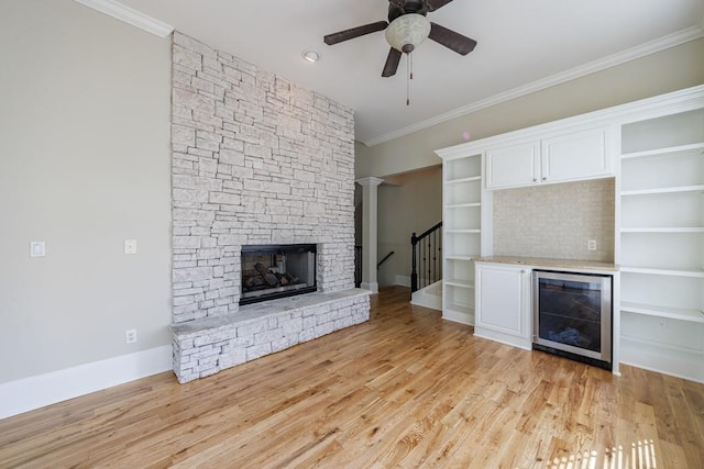 unfurnished living room with wine cooler, light wood-type flooring, a ceiling fan, crown molding, and a stone fireplace