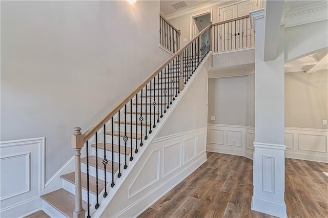 staircase featuring a decorative wall, wood finished floors, a towering ceiling, ornamental molding, and ornate columns