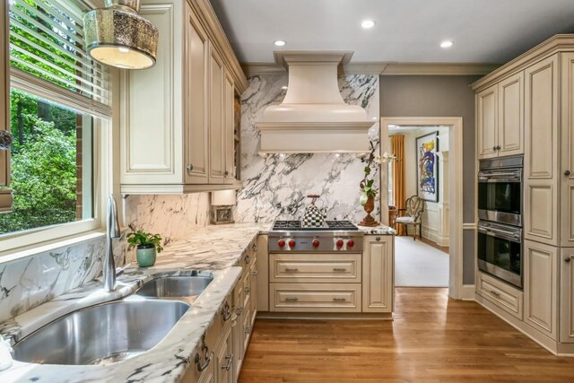 kitchen with decorative backsplash, cream cabinetry, custom range hood, a sink, and appliances with stainless steel finishes