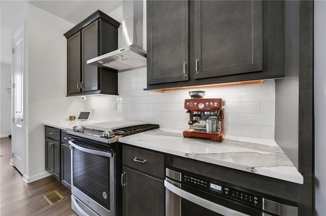 kitchen with light stone counters, dark wood-type flooring, appliances with stainless steel finishes, wall chimney range hood, and tasteful backsplash