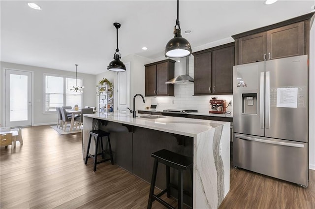 kitchen with a sink, dark brown cabinetry, wall chimney range hood, stainless steel fridge, and backsplash