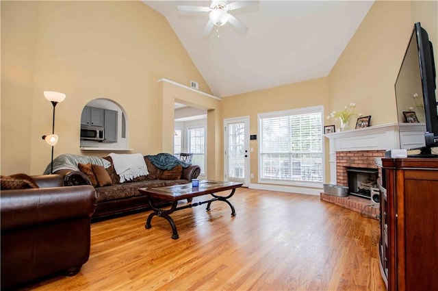 living area with ceiling fan, high vaulted ceiling, a brick fireplace, and light wood-style flooring