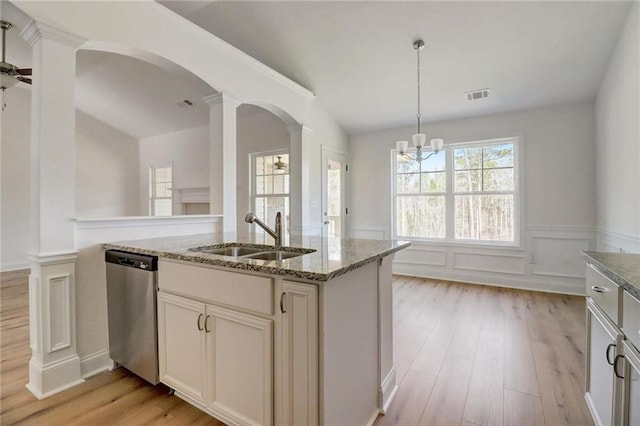 kitchen featuring sink, ornate columns, light stone counters, dishwasher, and an island with sink