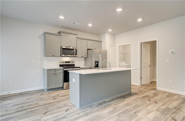 kitchen featuring sink, appliances with stainless steel finishes, gray cabinetry, a kitchen island with sink, and light wood-type flooring