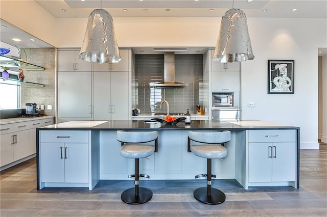 kitchen with tasteful backsplash, wall chimney exhaust hood, stainless steel microwave, wood finished floors, and a kitchen island with sink