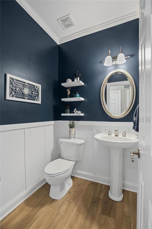 half bathroom with visible vents, ornamental molding, a decorative wall, wood finished floors, and a wainscoted wall