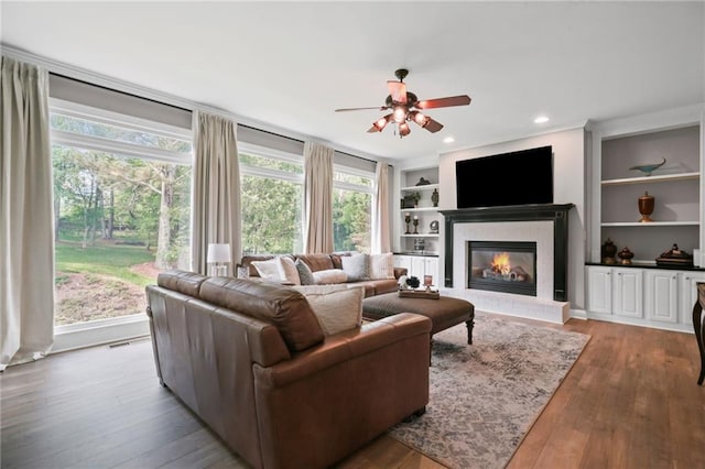 living room with ceiling fan, a glass covered fireplace, built in features, and wood finished floors