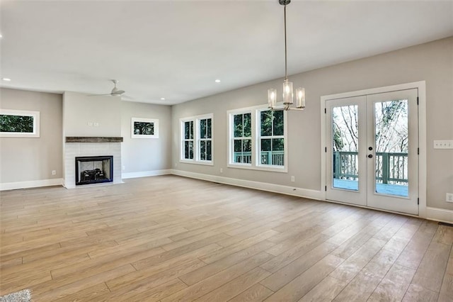 unfurnished living room with baseboards, a fireplace with flush hearth, french doors, light wood-style floors, and ceiling fan with notable chandelier