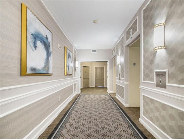 hallway with wainscoting, dark carpet, visible vents, and a decorative wall