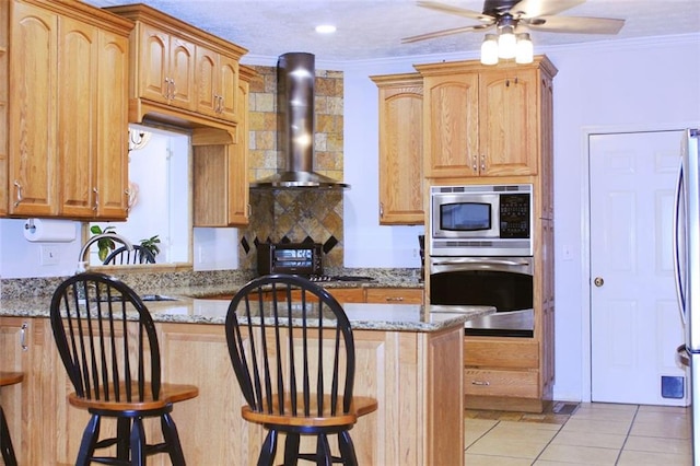 kitchen with light tile patterned floors, appliances with stainless steel finishes, ornamental molding, wall chimney range hood, and a kitchen breakfast bar