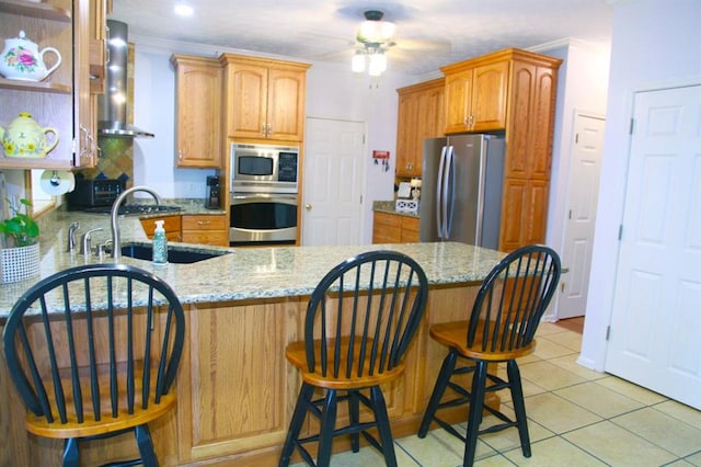 kitchen featuring light stone counters, appliances with stainless steel finishes, a sink, wall chimney range hood, and a peninsula