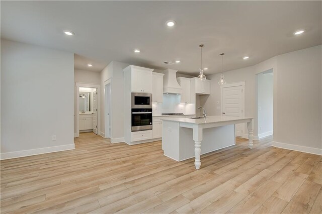 kitchen with premium range hood, a kitchen island with sink, light wood-type flooring, and appliances with stainless steel finishes