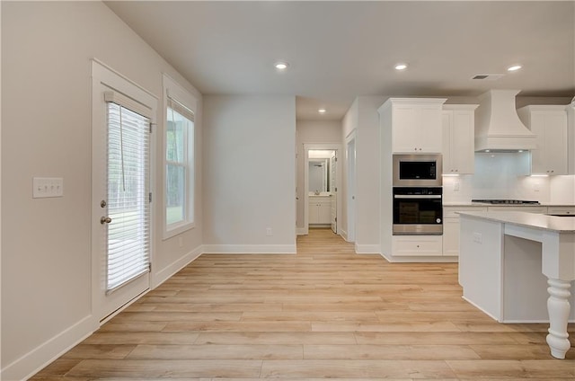 kitchen with white cabinetry, backsplash, light hardwood / wood-style floors, custom range hood, and appliances with stainless steel finishes