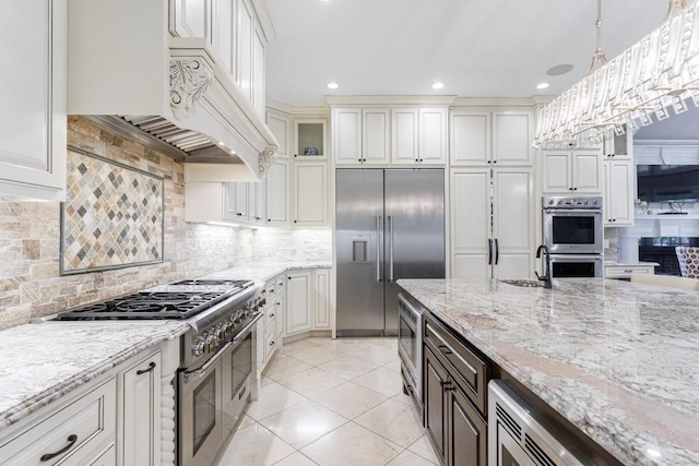 kitchen with custom range hood, decorative backsplash, hanging light fixtures, built in appliances, and white cabinetry