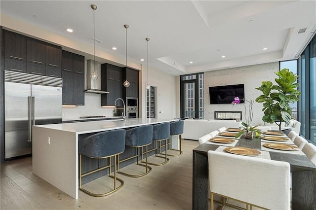 kitchen featuring wall chimney exhaust hood, stainless steel appliances, a center island with sink, and light hardwood / wood-style flooring