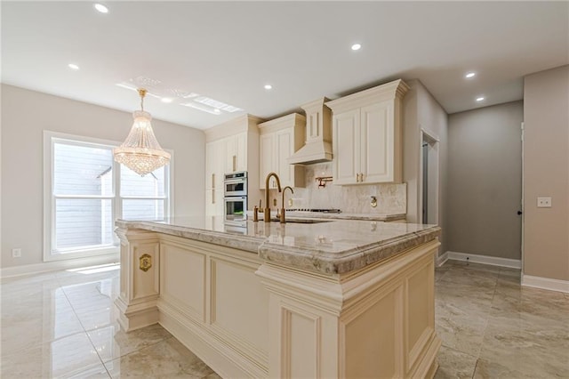 kitchen featuring custom range hood, stainless steel double oven, cream cabinets, a center island with sink, and decorative light fixtures