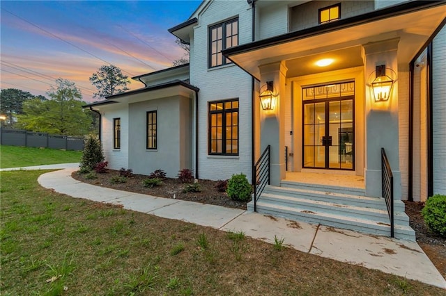 exterior entry at dusk with a lawn, brick siding, and fence