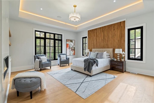 bedroom featuring a glass covered fireplace, light wood-style flooring, a raised ceiling, and baseboards