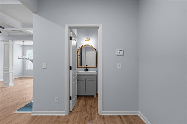 corridor featuring baseboards, decorative columns, light wood-style flooring, coffered ceiling, and a sink