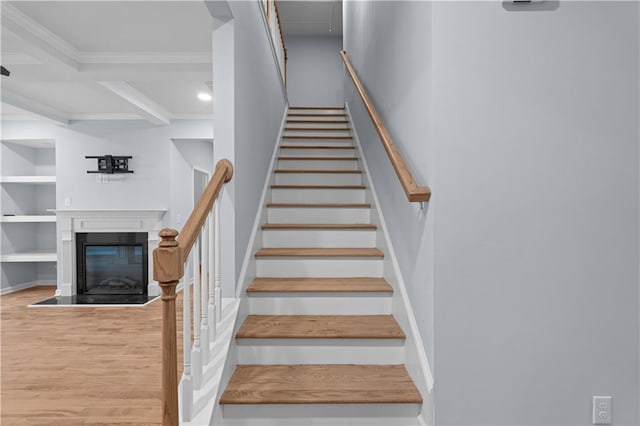 staircase featuring built in shelves, wood finished floors, coffered ceiling, a glass covered fireplace, and crown molding