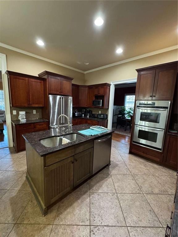 kitchen with sink, appliances with stainless steel finishes, dark stone countertops, ornamental molding, and a center island with sink