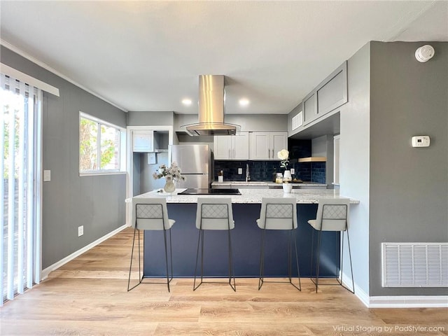 kitchen with kitchen peninsula, a kitchen bar, island range hood, stainless steel fridge, and light wood-type flooring