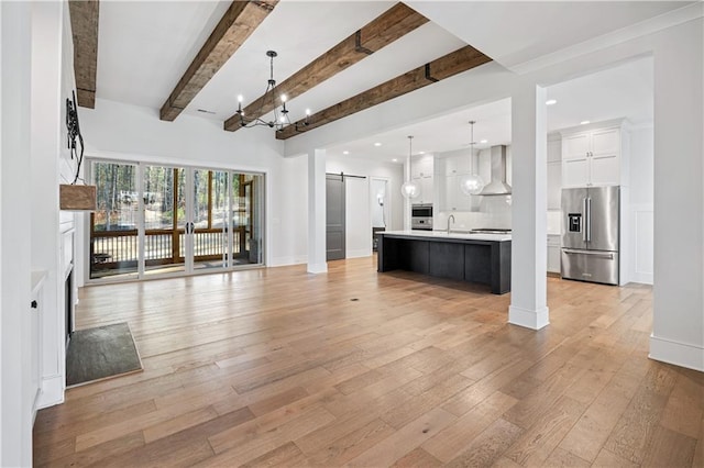 unfurnished living room featuring a barn door, light wood-style floors, a sink, a chandelier, and beamed ceiling